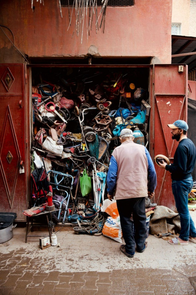 A man standing in front of an overfilled garage filled with tools, furniture, and scattered junk