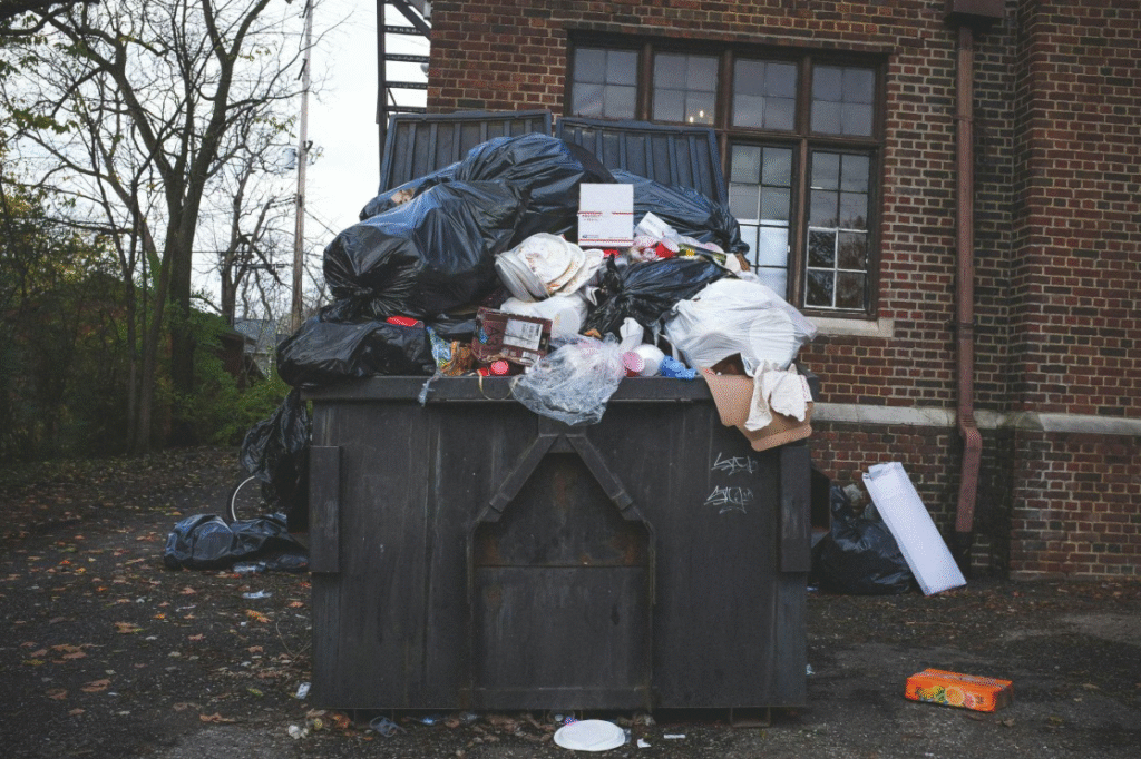 An overflowing black trash bin with piles of junk scattered across a residential property